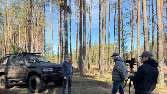 Pa kreisi silā redzams offroad džips, kas ir dubļains, pie tā stāv vīrietis, pa labi stāv 2 vīrieši, kur viens ir filmētājs, bet otrs intervētājs