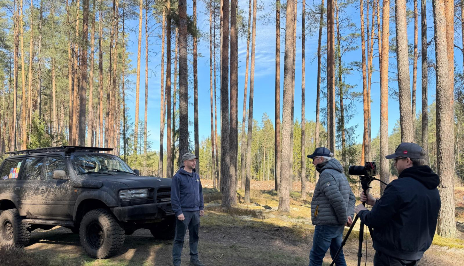 Pa kreisi silā redzams offroad džips, kas ir dubļains, pie tā stāv vīrietis, pa labi stāv 2 vīrieši, kur viens ir filmētājs, bet otrs intervētājs